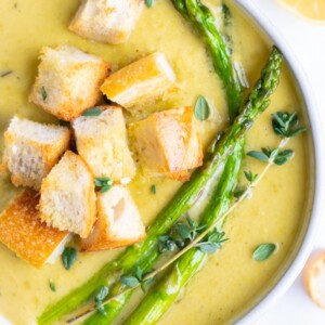 Cream of asparagus soup is in a white bowl with croutons and asparagus spears.