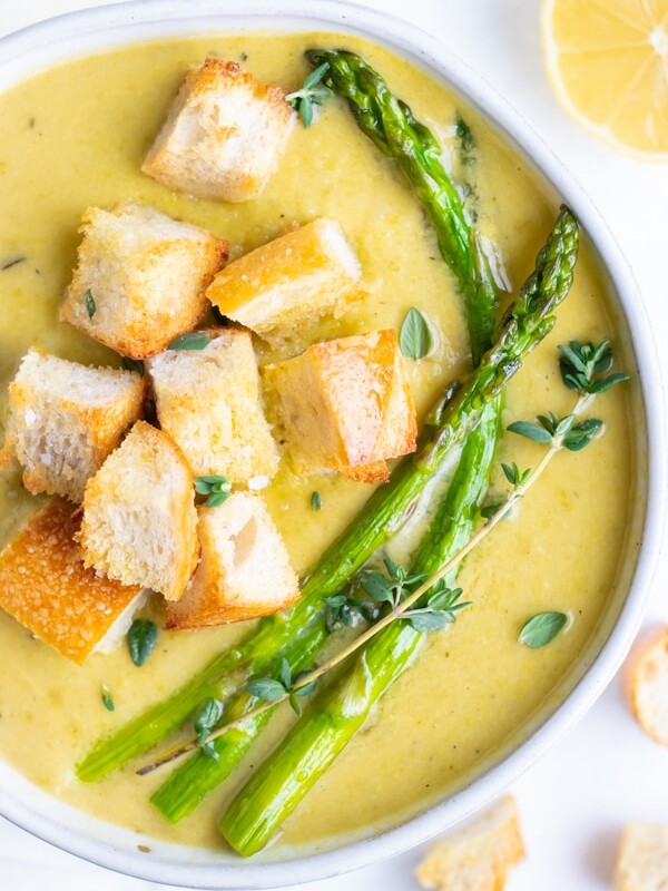Cream of asparagus soup is in a white bowl with croutons and asparagus spears.