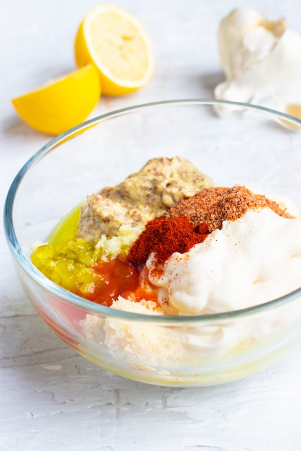 A clear bowl with mayonnaise, mustard, paprika, Cajun seasoning, and pickle relish with lemons and garlic in the background.