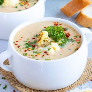 A white soup bowl full of cream of cauliflower soup with cauliflower florets and parsley on top.