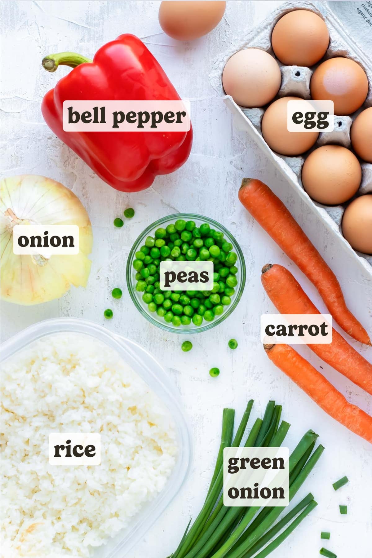 Eggs, rice, red bell pepper, onion, carrots, and green onion arranged on a white countertop.