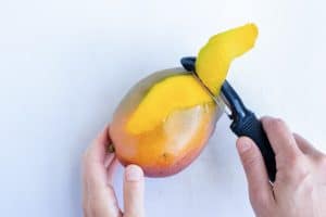 A black vegetable peeler removes the skin from a mango.