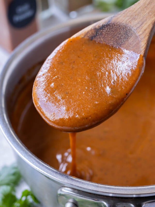 A wooden spoon is scooping up a homemade red enchilada sauce from a saucepan.