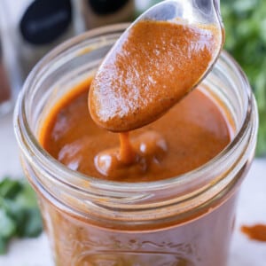 A homemade red enchilada sauce is being scooped out of a glass jar with a spoon.