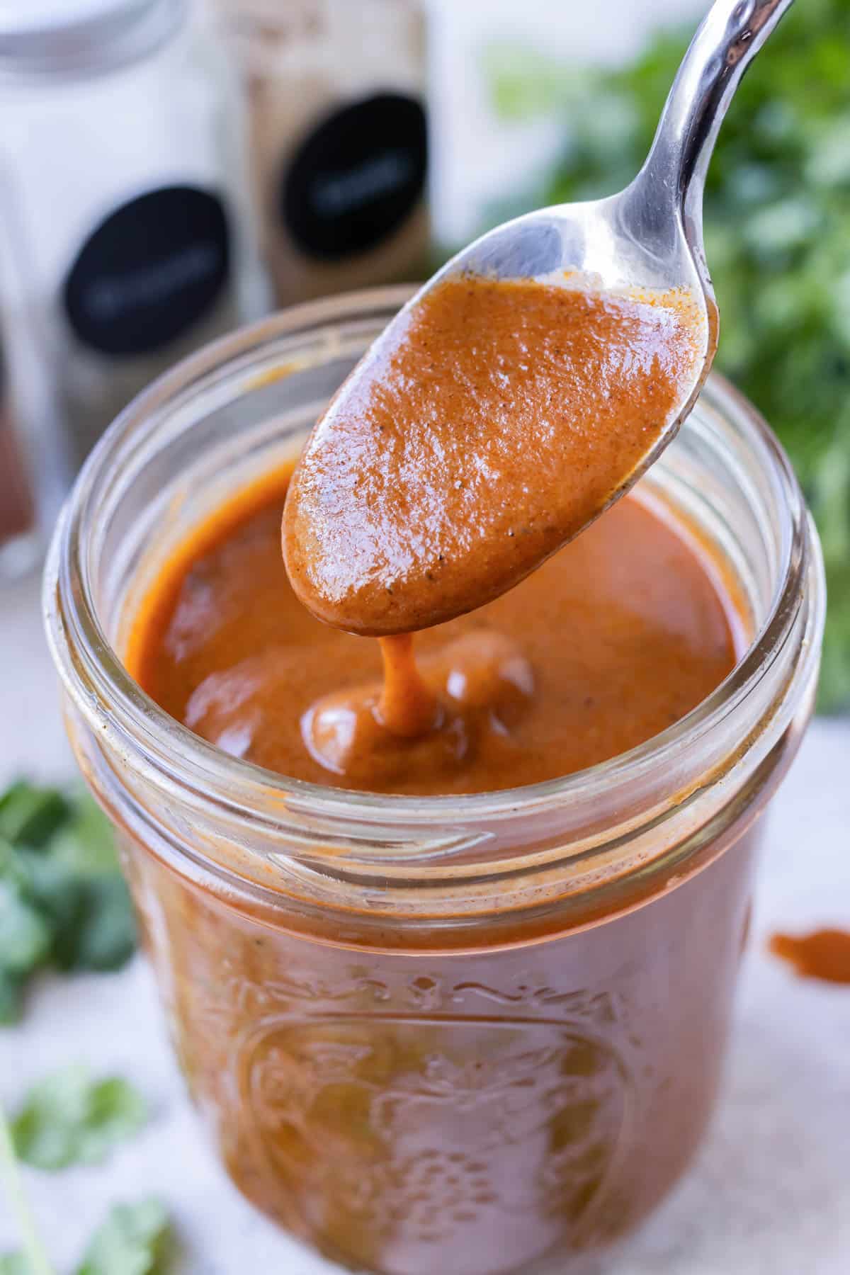 A homemade red enchilada sauce is being scooped out of a glass jar with a spoon.