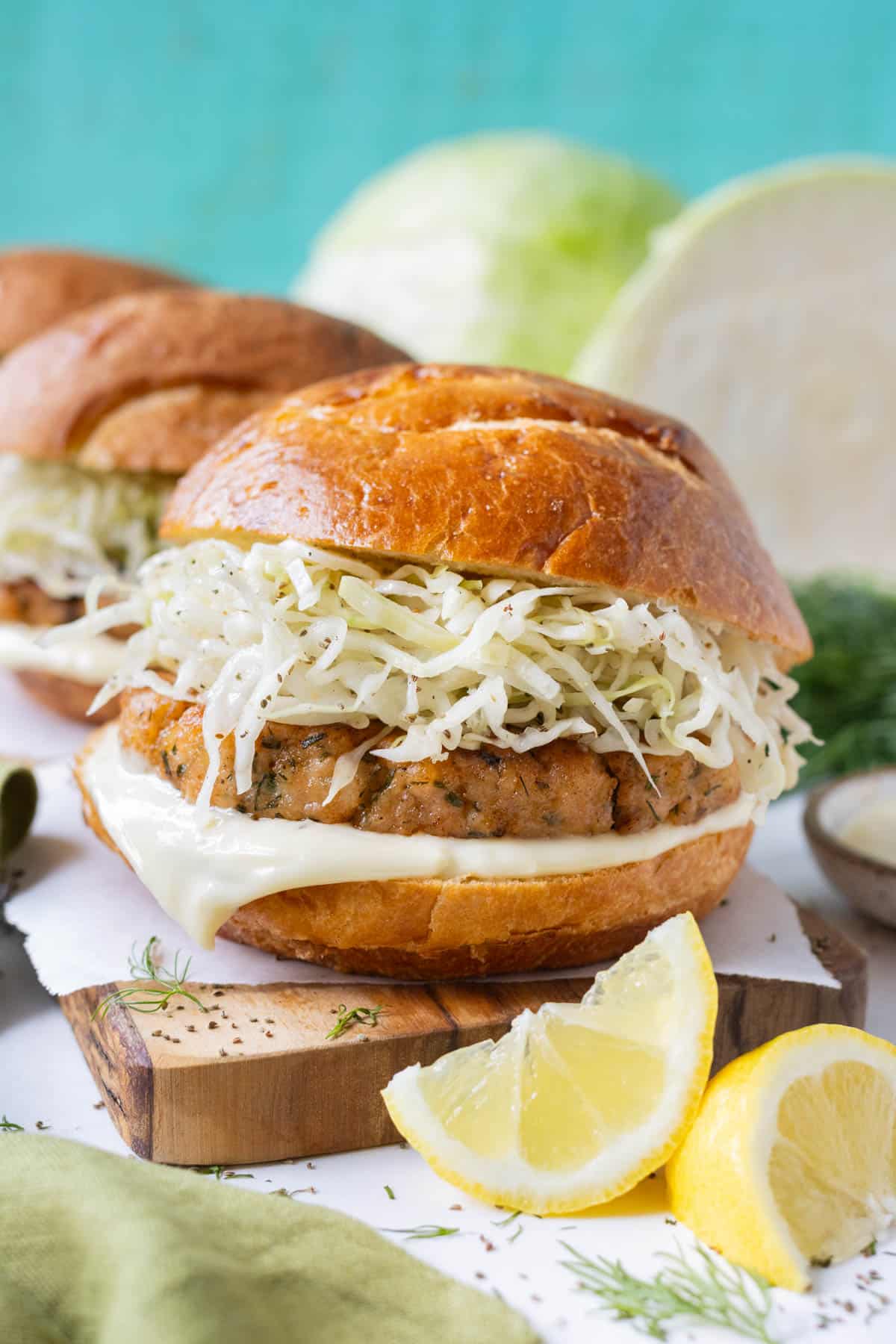 Salmon burger patties with a vinegar-based coleslaw on toasted sweet buns with lemon wedges next to it.