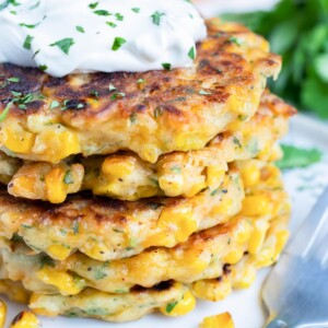 Fried corn fritters are in a stack on a white plate with a dollop of sour cream on top.