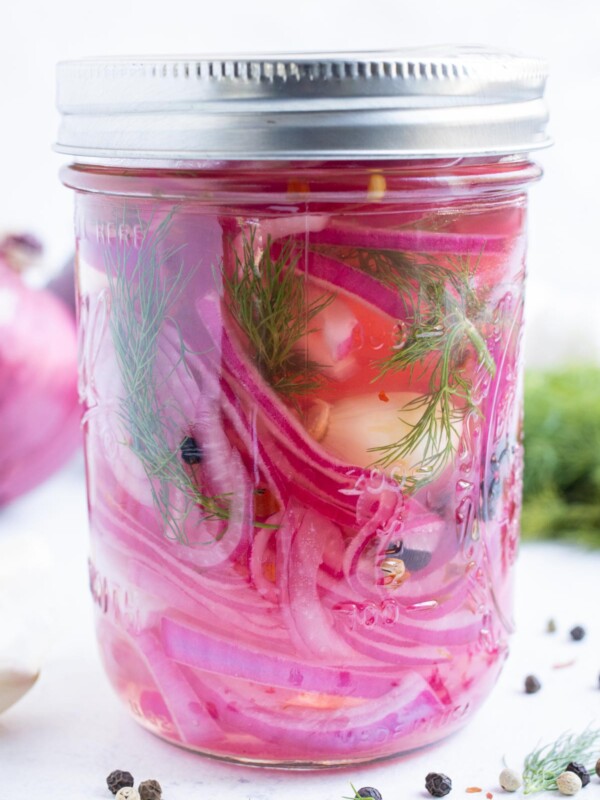 A closeup of red onions, garlic, and herbs in a jar filled with a brining solution.