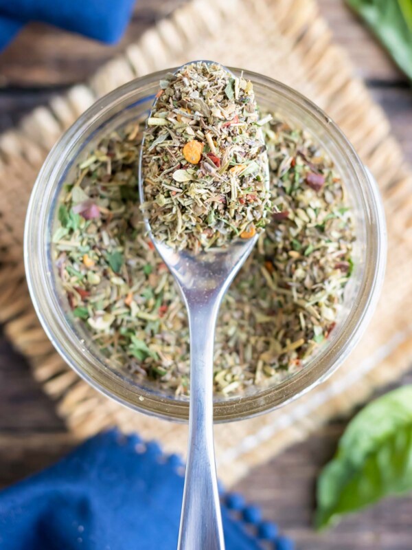 A clear glass jar with a spoon scooping out a homemade Italian seasoning mix.