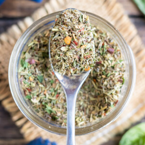 A clear glass jar with a spoon scooping out a homemade Italian seasoning mix.