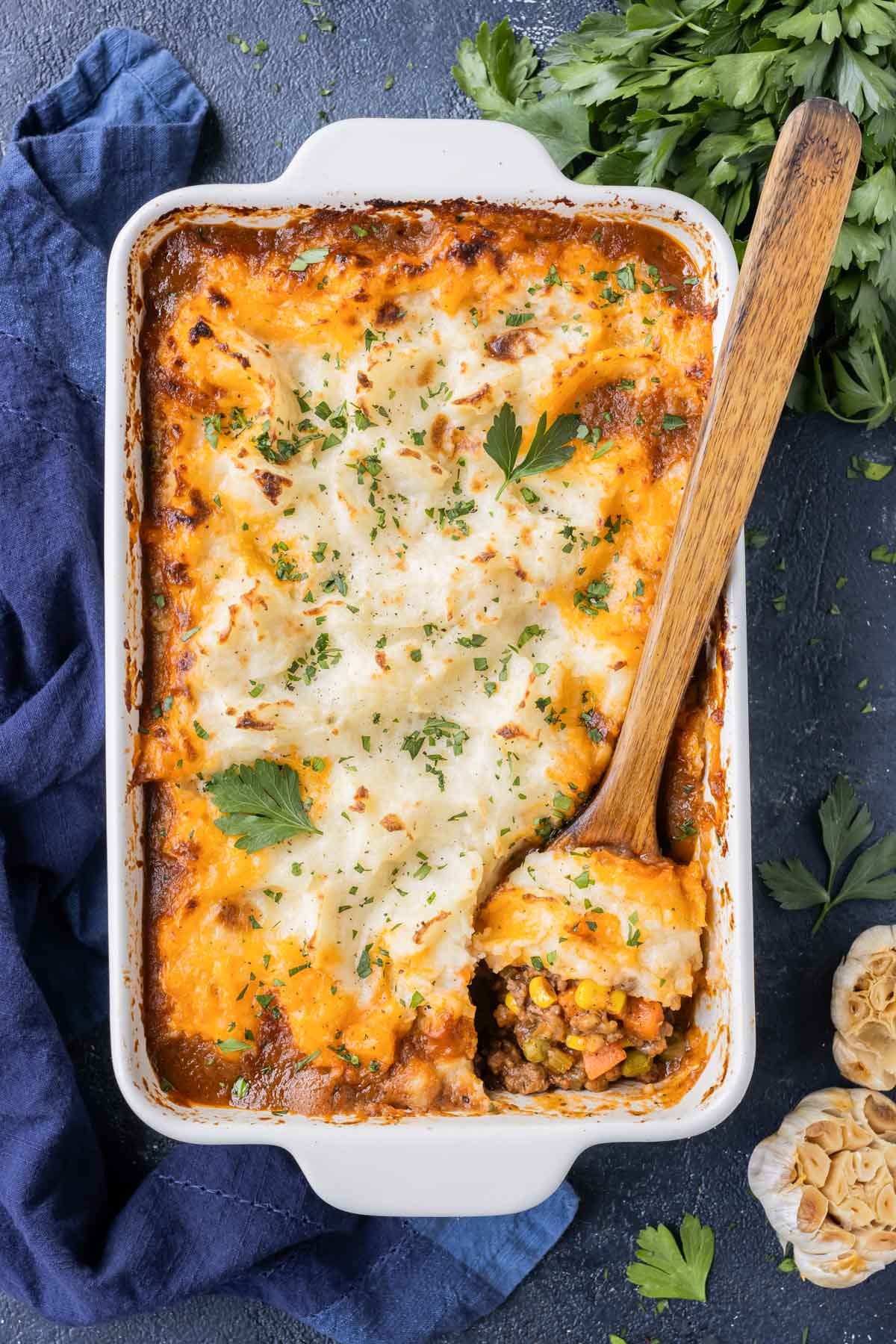 A traditional shepherd's pie recipe in a white baking dish with a wooden spoon scooping up a serving.