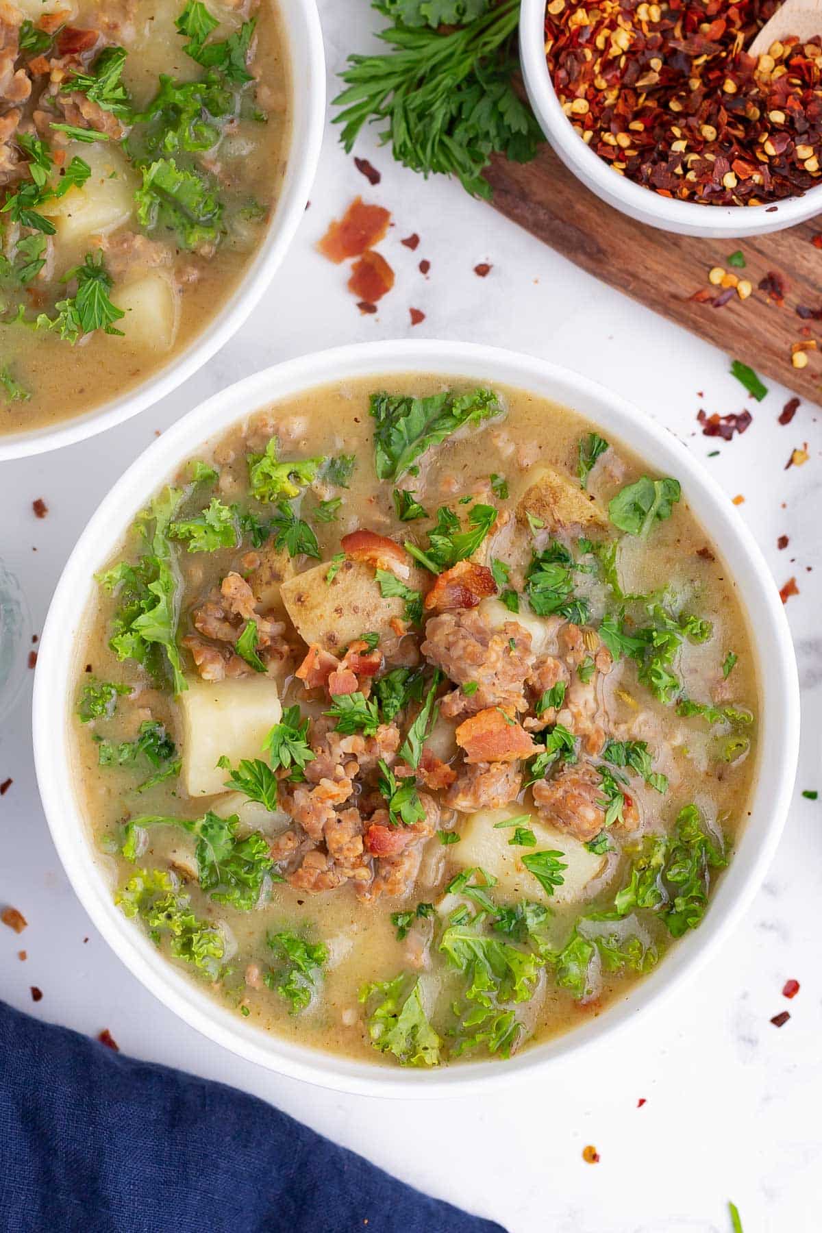 Two bowls full of zuppa toscana soup made from a copycat Olive Garden recipe are on a counter with bacon, potatoes, and Italian sausage.