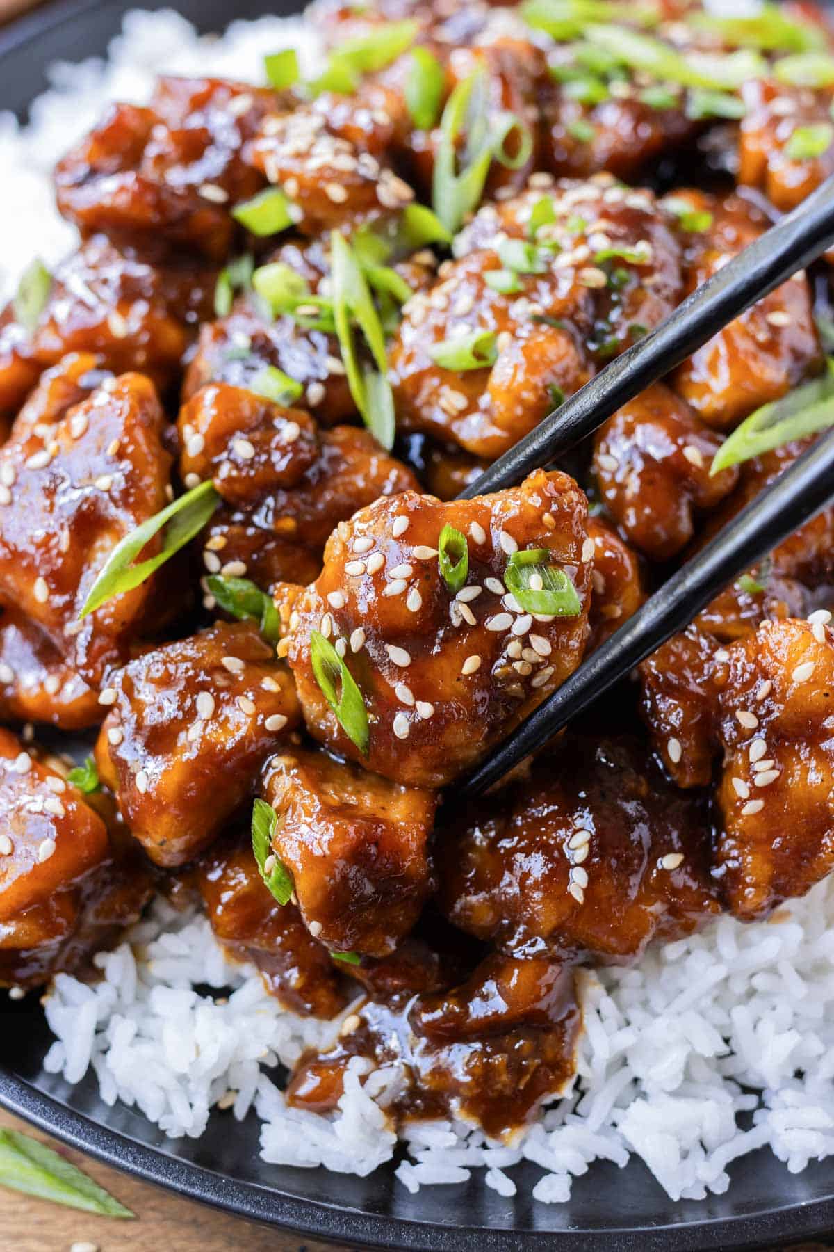 A piece of General Tso's chicken is being picked up with chopsticks from a bowl.