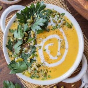 A bowl full of roasted butternut squash soup with coconut milk that's garnished with pumpkin seeds and parsley leaves.