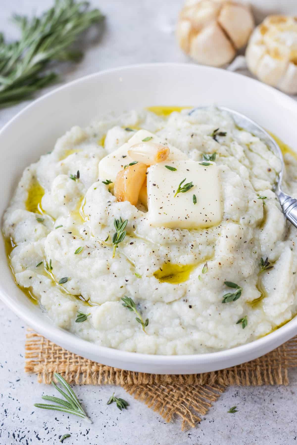 Garlic mashed cauliflower is served in a white bowl with cloves of roasted garlic, thyme, and rosemary on top.