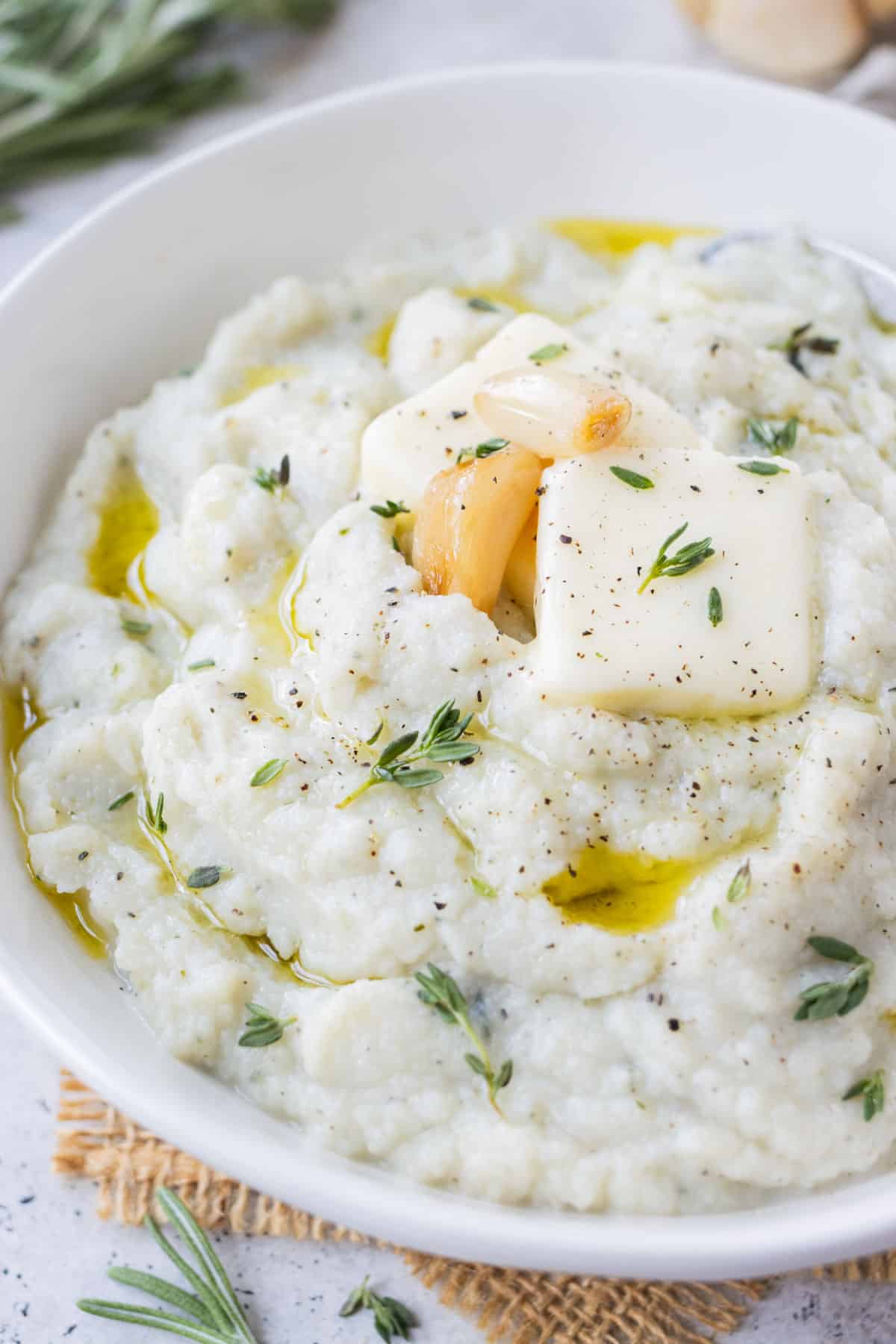 Garlic mashed cauliflower is served in a white bowl with cloves of roasted garlic, thyme, and rosemary on top.