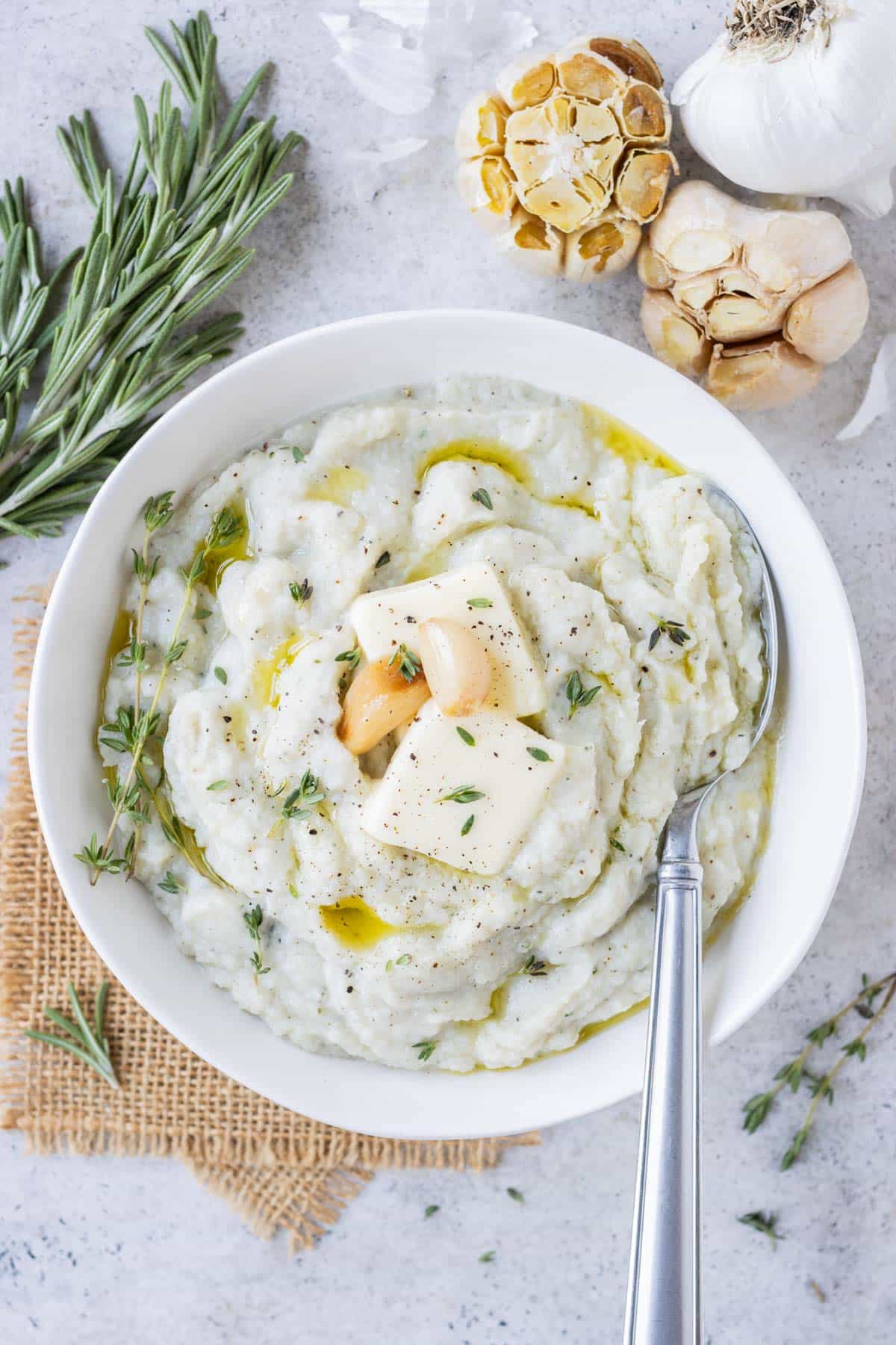 Garlic mashed cauliflower is served in a white bowl with a spoon and cloves of roasted garlic, thyme, and rosemary on top.