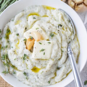 Garlic mashed cauliflower is served in a white bowl with a spoon and cloves of roasted garlic, thyme, and rosemary on top.