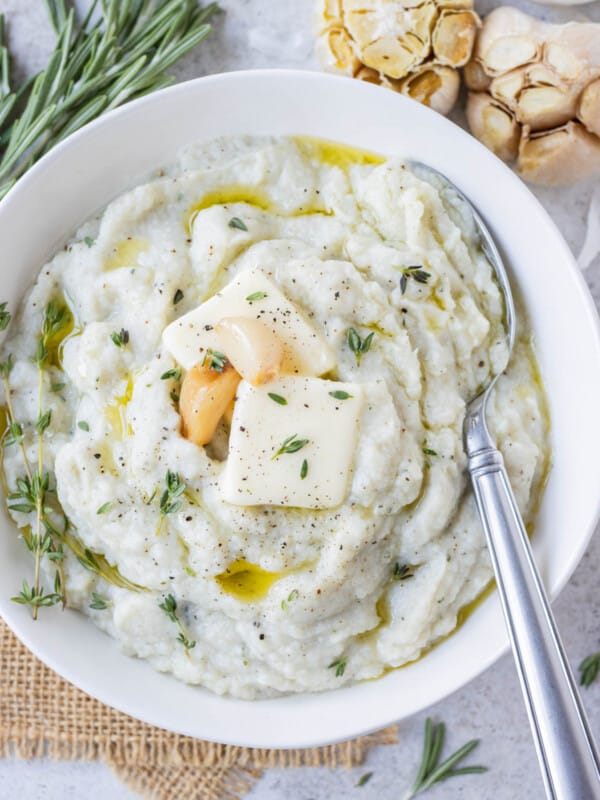 Garlic mashed cauliflower is served in a white bowl with a spoon and cloves of roasted garlic, thyme, and rosemary on top.