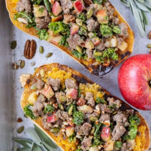 Two butternut squash halves are stuffed with ground turkey, kale, and pecans in a metal pan with an apple.