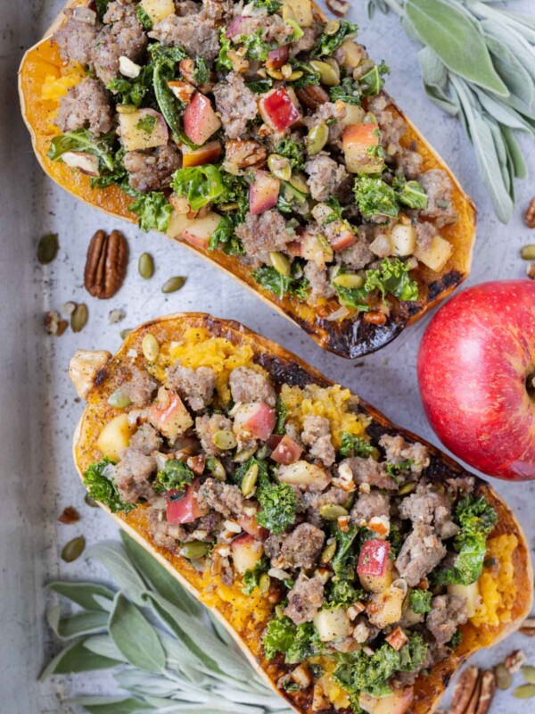 Two butternut squash halves are stuffed with ground turkey, kale, and pecans in a metal pan with an apple.