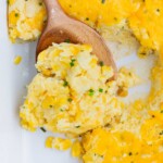 A wooden spoon scoops a serving of corn casserole from a pan.