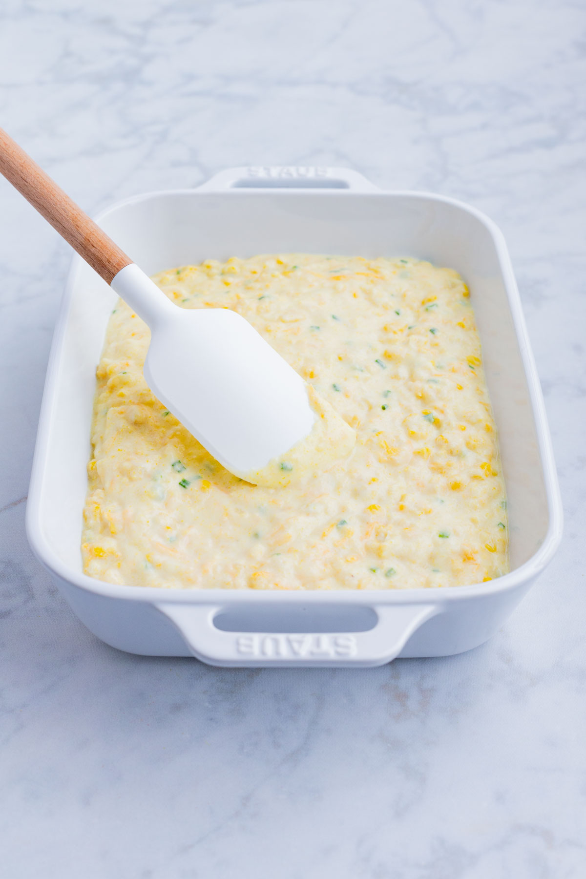 The corn casserole batter is spread in a white baking dish.