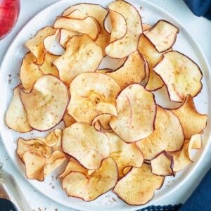 Air fryer apple chips are on a white plate next to apples and cinnamon.