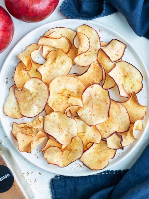 Air fryer apple chips are on a white plate next to apples and cinnamon.