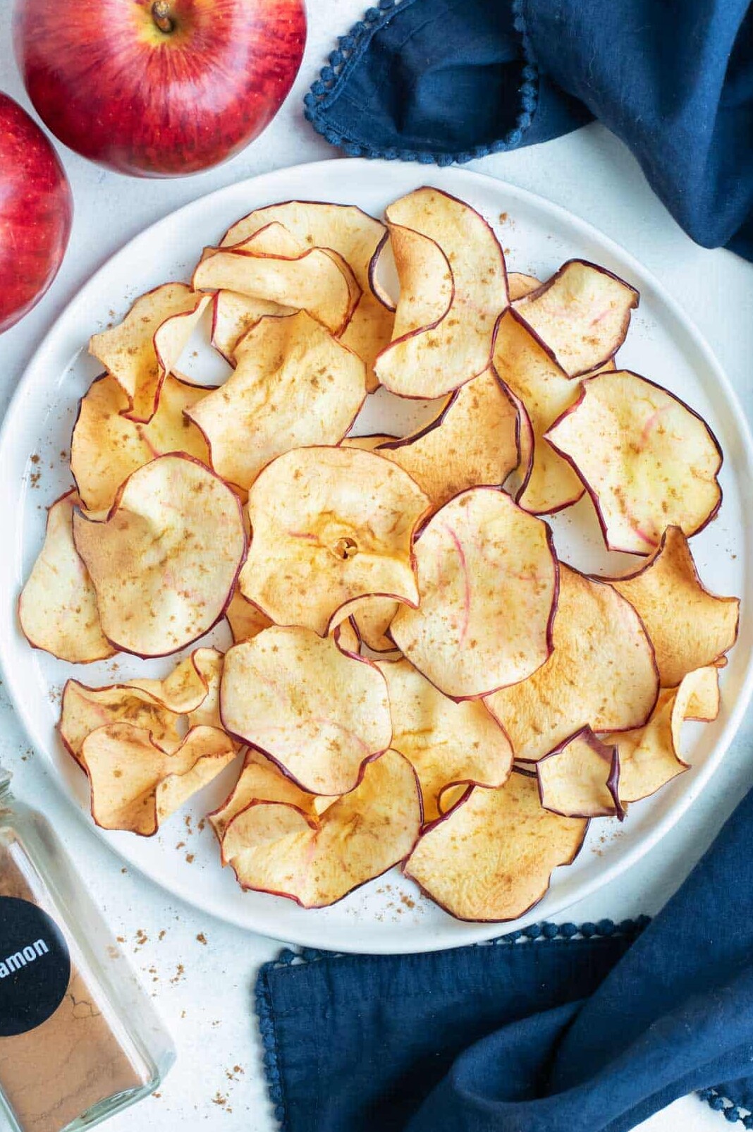 Air fryer apple chips are on a white plate next to apples and cinnamon.