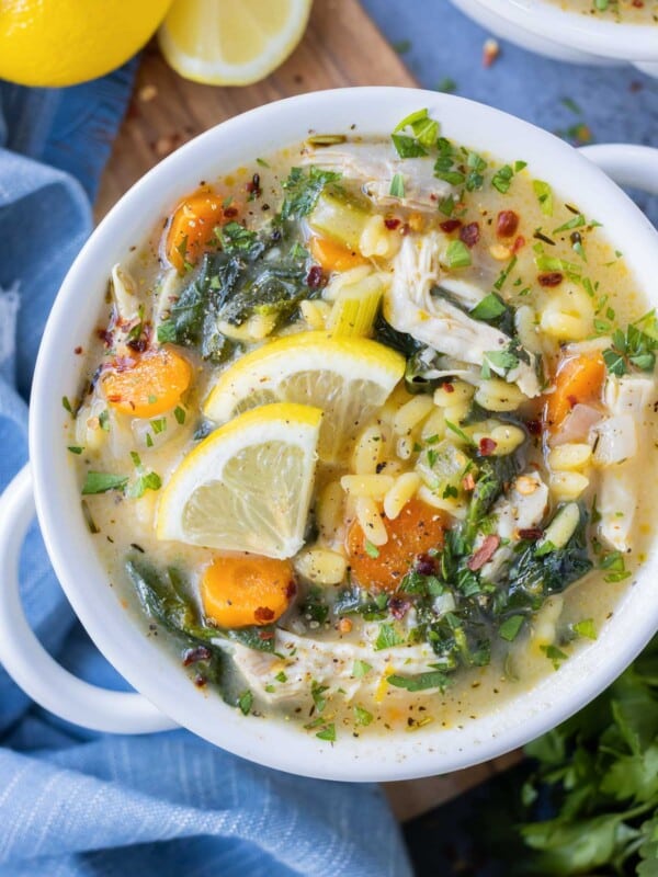 A bowl full of Lemon Chicken Orzo Soup filled with orzo pasta, spinach, carrots, and lemon slices is on a wooden board.