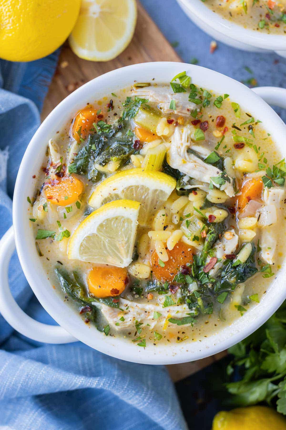 A bowl full of Lemon Chicken Orzo Soup filled with orzo pasta, spinach, carrots, and lemon slices is on a wooden board.