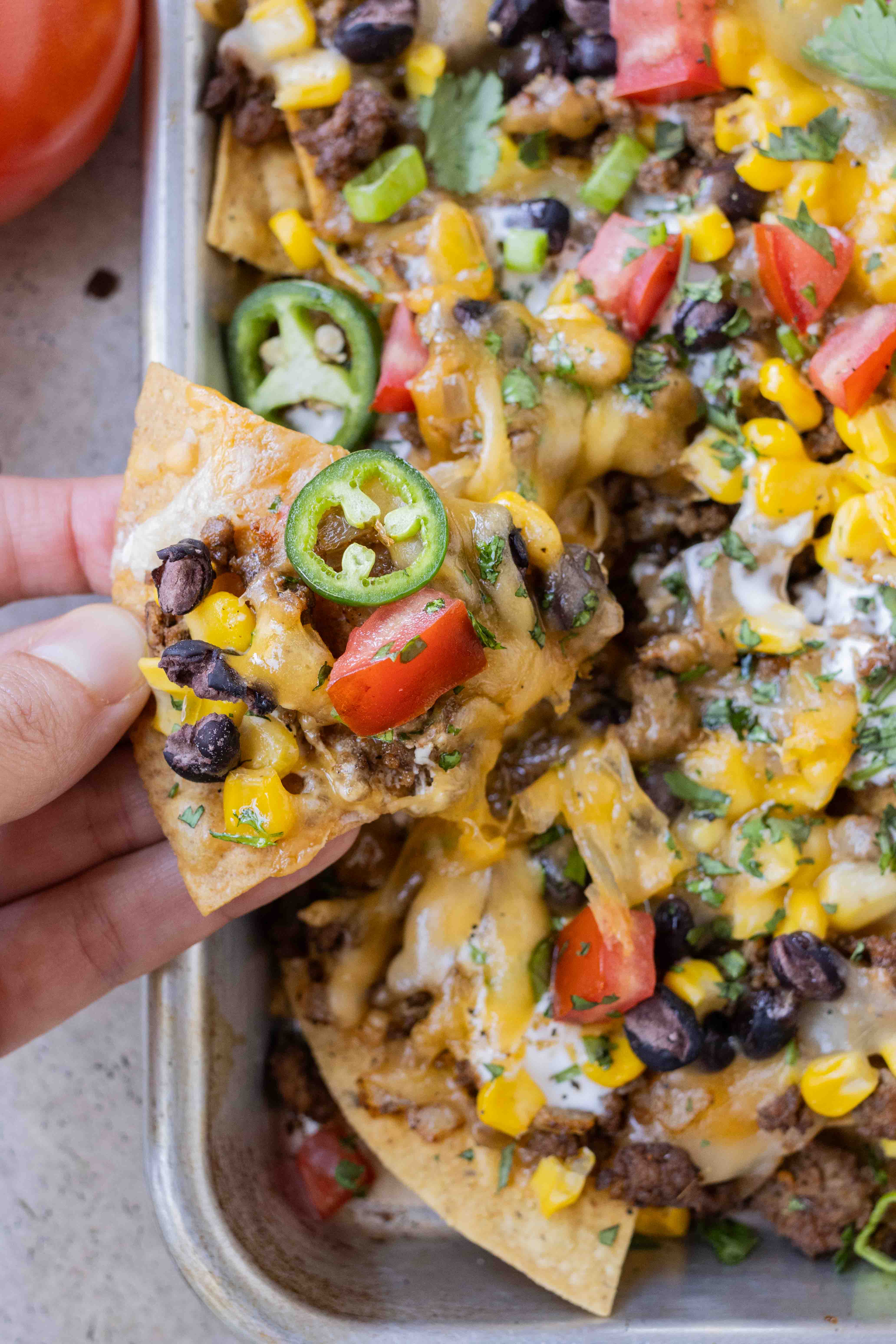 A hand picks up a chip loaded with toppings from a tray of sheet pan nachos.