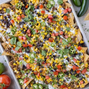 Sheet pan loaded nachos with ground beef, corn, cheese, and beans for an easy weeknight dinner or game day appetizer are next to tomatoes and jalapenos.