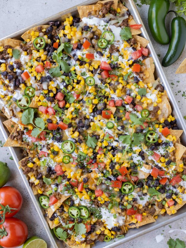 Sheet pan loaded nachos with ground beef, corn, cheese, and beans for an easy weeknight dinner or game day appetizer are next to tomatoes and jalapenos.