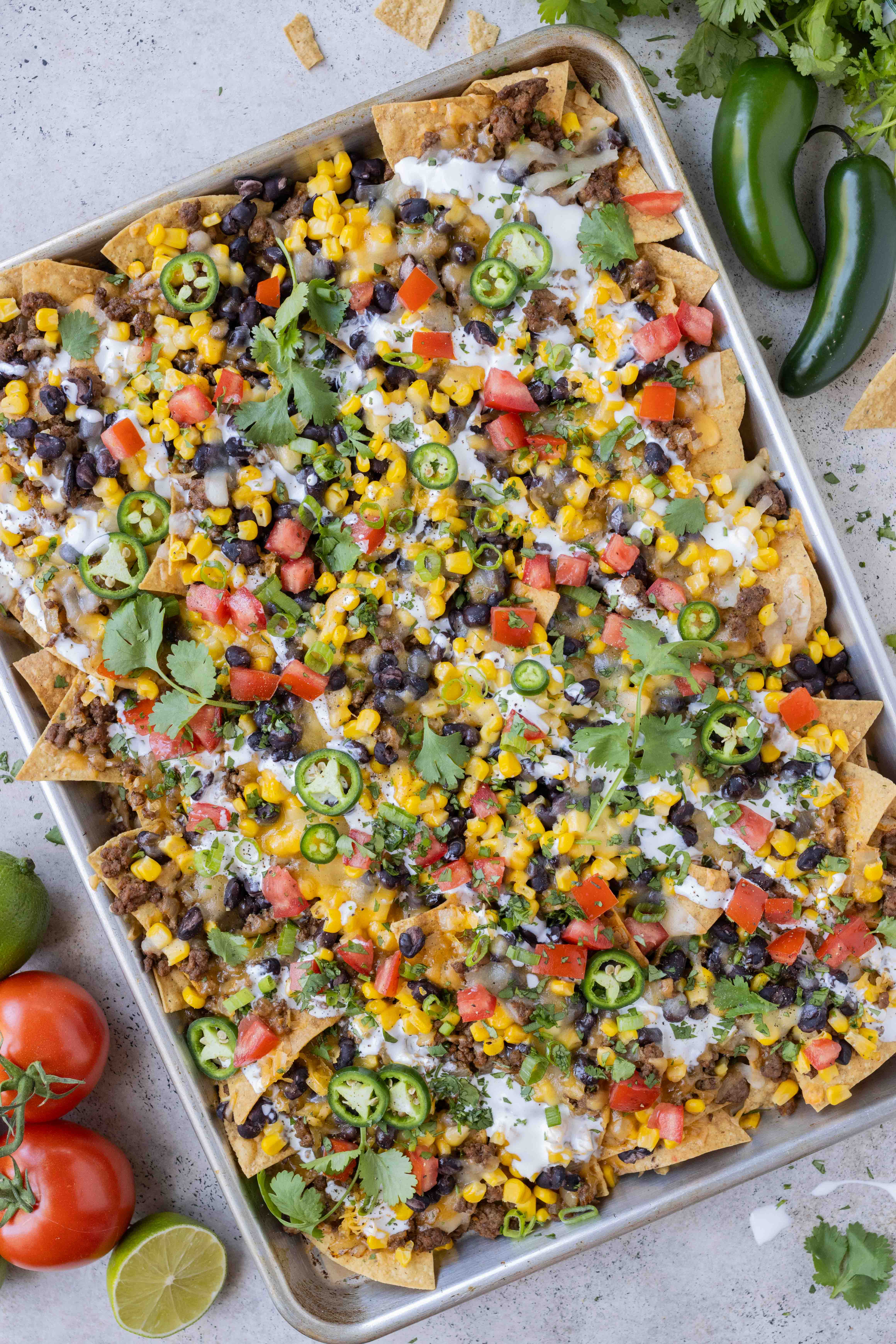 Sheet pan loaded nachos with ground beef, corn, cheese, and beans for an easy weeknight dinner or game day appetizer are next to tomatoes and jalapenos.