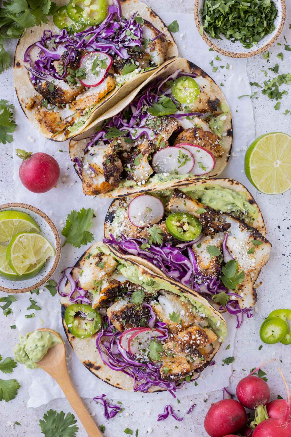 A row of tacos full of blackened fish, avocado cream sauce, and toppings are served on a sheet of wax paper.