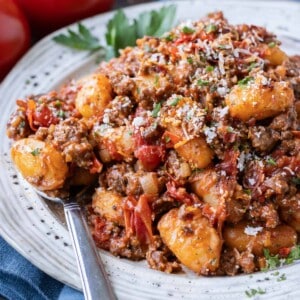 Gnocchi beef bolognese is served on a plate with a spoon and topped with Parmesan cheese.