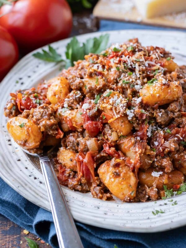 Gnocchi beef bolognese is served on a plate with a spoon and topped with Parmesan cheese.