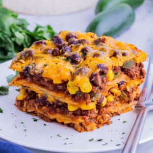 A slice of taco lasagna with corn tortillas is on a white plate with a fork next to it.