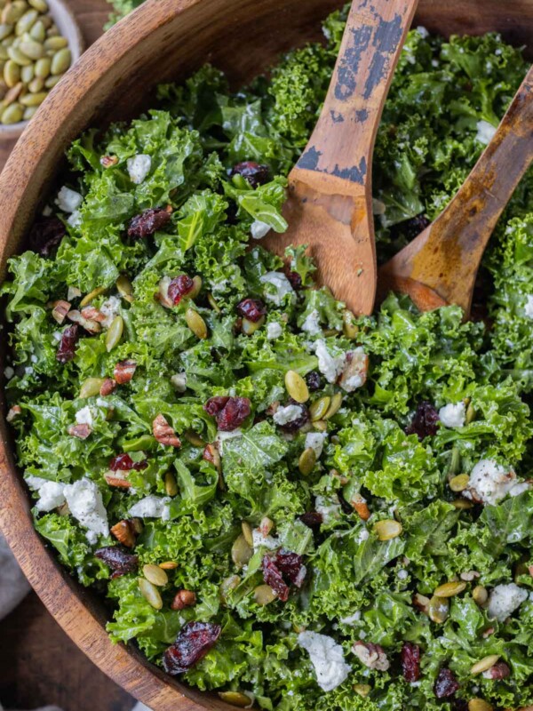 A chopped kale salad with cranberries, goat cheese, pecans, and pumpkin seeds in a wooden bowl with serving spoons for a Fall dinner.