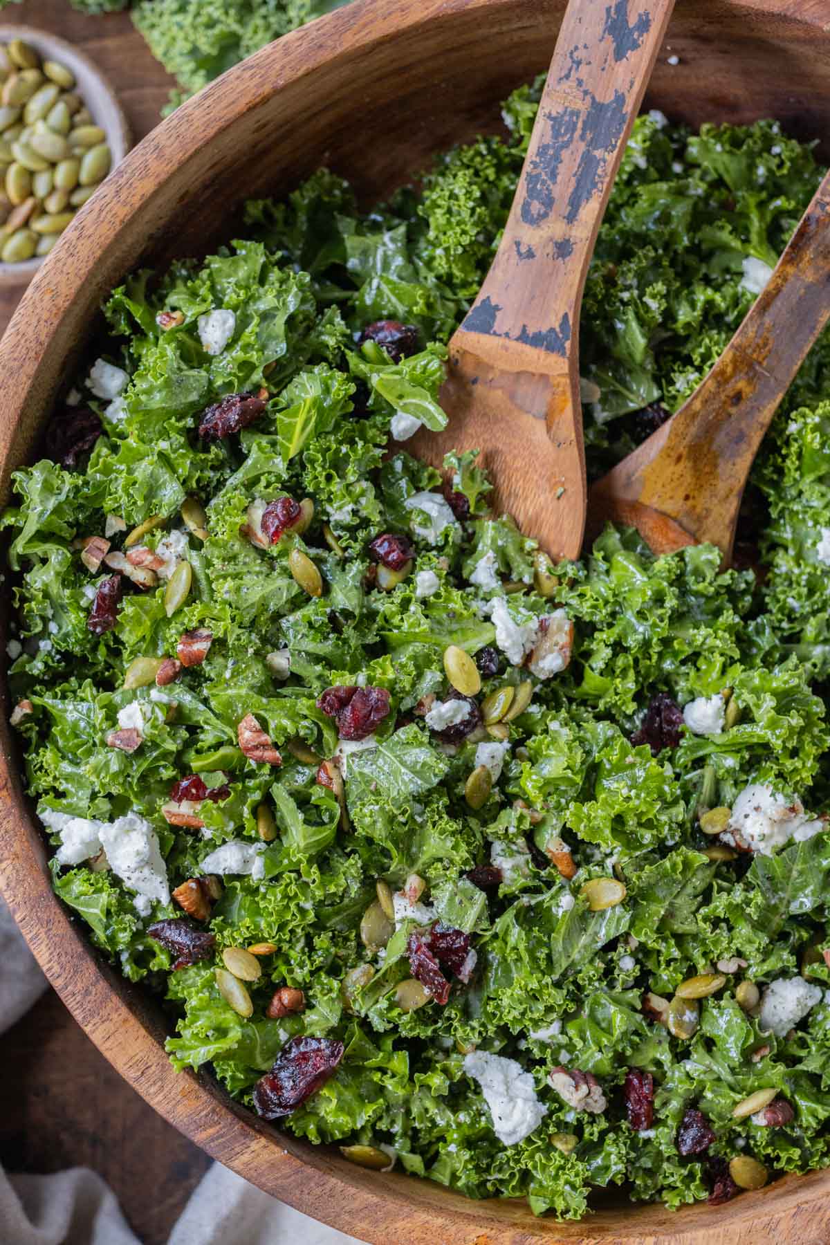 A chopped kale salad with cranberries, goat cheese, pecans, and pumpkin seeds in a wooden bowl with serving spoons for a Fall dinner.