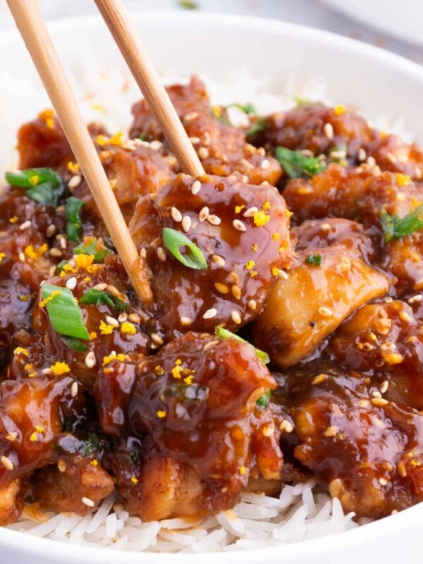 Chopsticks pick up a single piece of orange chicken from a bowl with rice.