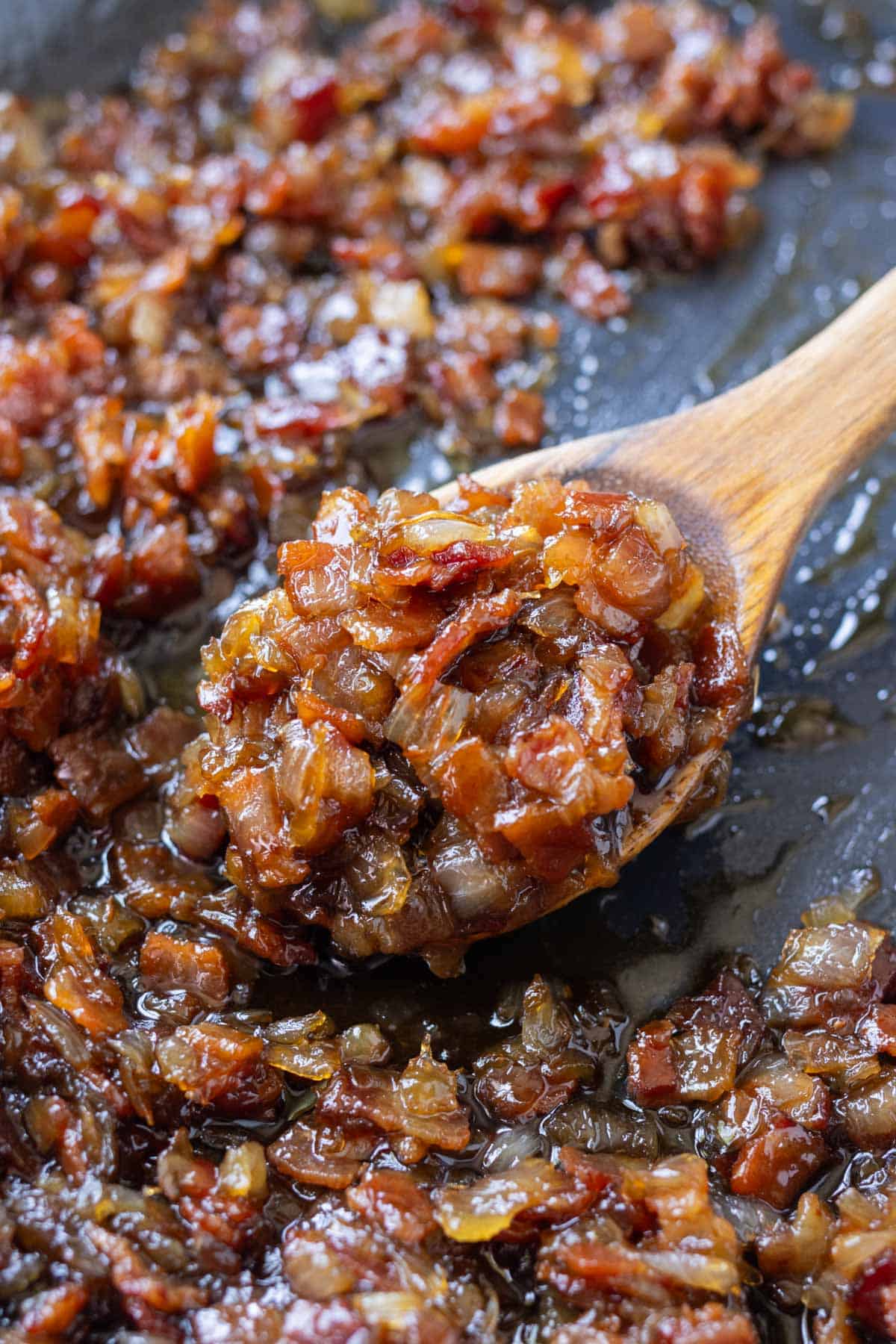 A wooden spoons scoops up some bacon jam with onions from a skillet.