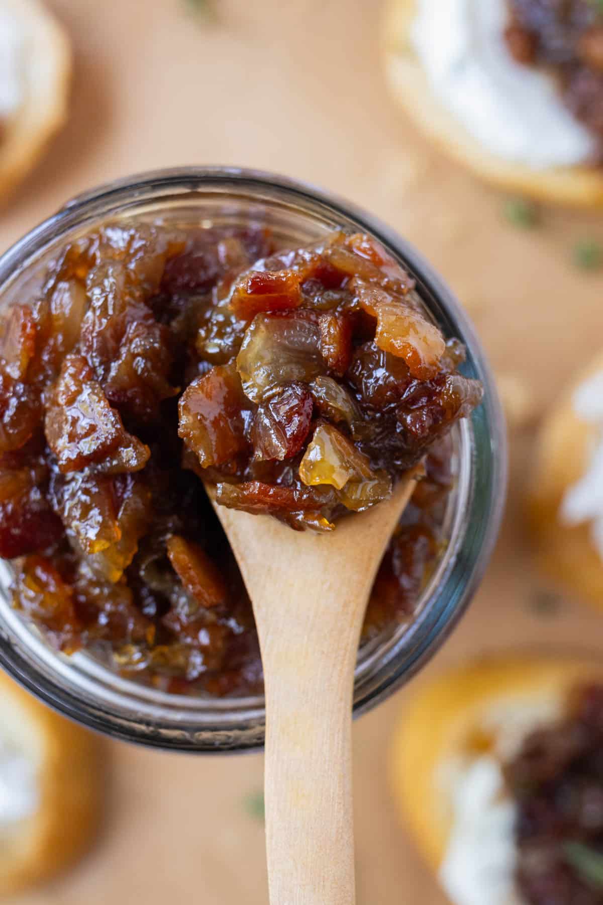A wooden spoons scoops up some bacon onion jam from a glass jar.