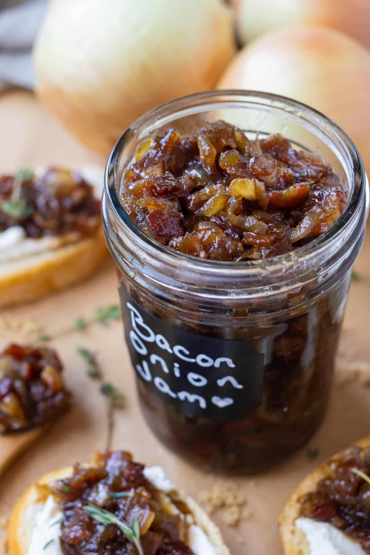 Balsamic bacon onion jam is stored in a glass mason jar with crostinis surrounding it.