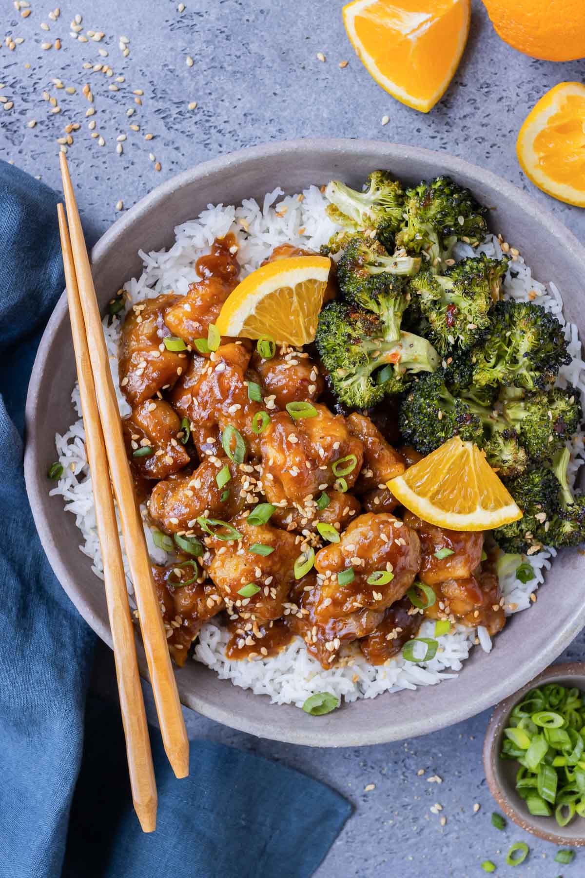 Crispy orange chicken is in a bowl full of rice and served with roasted broccoli with fresh orange wedges on the side.