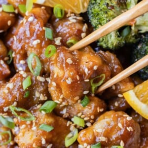 Chopsticks are picking up a single piece of orange chicken from a bowl.