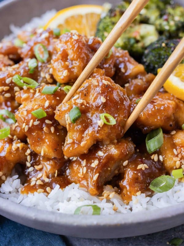 Chopsticks are picking up a piece of crispy orange chicken from a bowl filled with rice and broccoli.