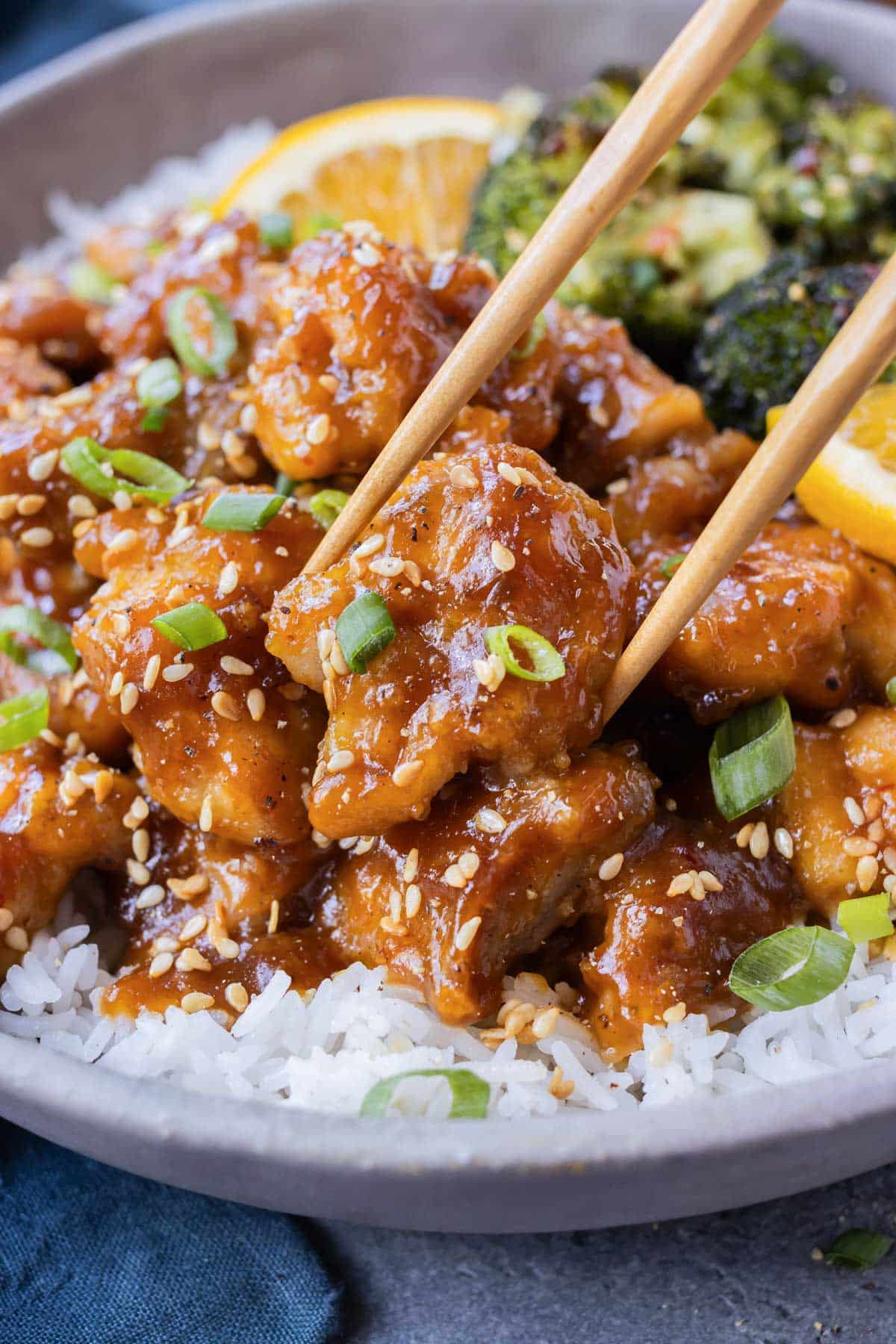 Chopsticks are picking up a piece of crispy orange chicken from a bowl filled with rice and broccoli.